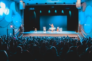An auditorium full of people watch a performer on a stage as in a play.